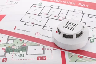 Smoke detector placed on top of office fire evacuation floor plans with labeled exits, alarm buttons, and fire extinguishers.